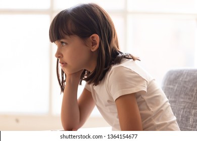 Side profile view brunette little girl adorable daughter sitting alone at home near window head rests on the hand preschool kid thinking dreaming imagining. Pose of boredom disinterest fatigue conceptの写真素材