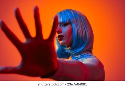 Side view of confident young woman in blue wig with red lips covering camera with hand against orange background 库存照片