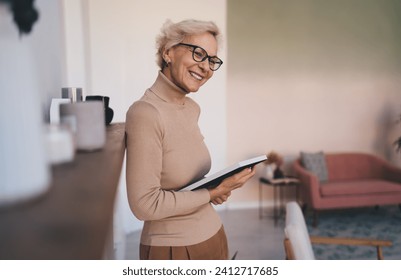 Side view of aged smiling lady in casual wear and spectacles holding photo frame and reminiscing while standing near shelf in living room Stock fotografie