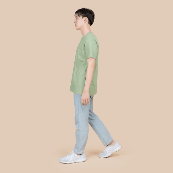 Vista lateral de un joven asiático a media zancada, caminando despreocupadamente. Lleva una camiseta verde, unos vaqueros azules y unas zapatillas blancas sobre un fondo beige neutro Foto de stock