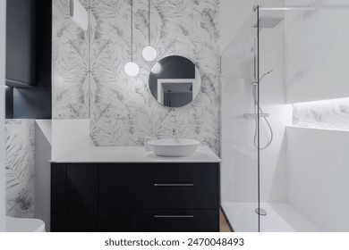 A sleek modern bathroom featuring a stylish vanity with a round mirror, glass shower enclosure, and elegant lighting. The sophisticated design includes marble-patterned walls and contemporary fixtures Arkistovalokuva