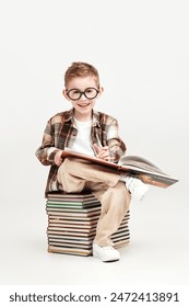 Un Pequeño y alegre chico inteligente con gafas se sienta en una pila de libros y sonríe felizmente. Fondo de estudio blanco. Educación y desarrollo temprano de los niños.  Foto de stock