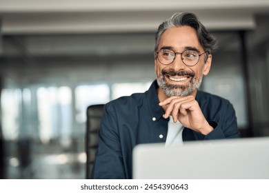Smiling mature business man executive wearing shirt sitting at desk using laptop. Happy busy professional middle aged Indian businessman investor working on computer looking away in office. Copy space Foto stock