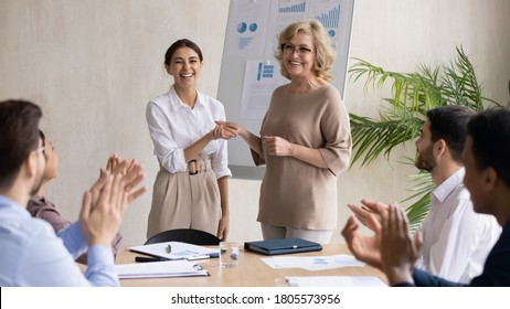 Smiling middle aged 60s female company leader shaking hands with happy young arabic colleague, praising for good work or thanking for good job done at briefing meeting with diverse employees. Arkistovalokuva