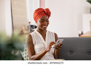 Smiling middle aged african woman with traditional head turban sitting on couch at home using smartphone. Beautiful african american woman with typical headscarf scrolling through internet on phone., fotografie de stoc