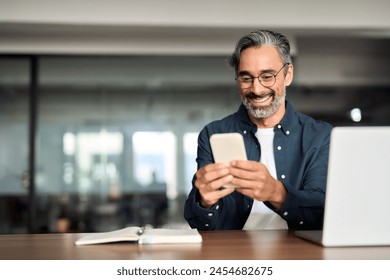 Smiling older Indian business man sitting at desk using mobile phone. Happy mature businessman executive, busy middle aged entrepreneur investor looking at smartphone working in office with cellphone. Stockfoto