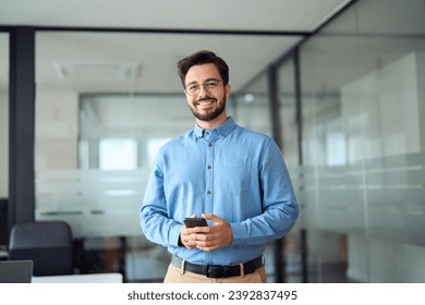 Smiling busy professional latin business man standing in office holding mobile cellphone. Young happy businessman employee using smartphone looking at camera using cell phone tech at work. Portrait. Foto stock