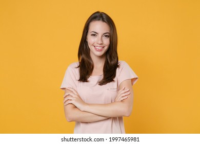 Sonriente hermosa y atractiva joven morena de 20 años con camiseta rosa pastel casual posando con las manos cruzadas cámara aislada en un brillante retrato de estudio de fondo de pared de color amarillo Foto de stock