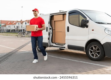 Smiling delivery man in uniform carries package from his white van, delivering parcel to customer, walking outdoors 库存照片