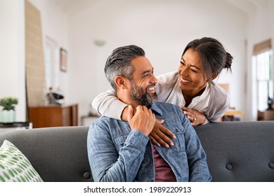 Smiling ethnic woman hugging her husband on the couch from behind in the living room. Middle eastern man having fun with his beautiful young wife on the couch. Mid adult indian man with latin woman., fotografie de stoc