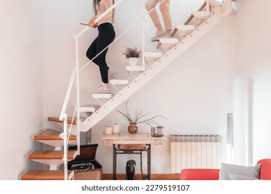 Smiling female with tablet and unrecognizable lady climbing down steps while holding white railing and staircase decorated with plant retro record player table with marble pots in living room Stock Photo