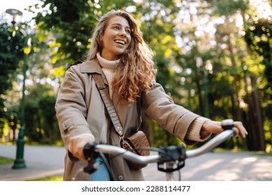 Smiling woman with curly hair in a coat rides a bicycle in a sunny park. Outdoor portrait. Beautiful woman enjoys nature. Lifestyle. Relax, nature concept.  – Ảnh có sẵn