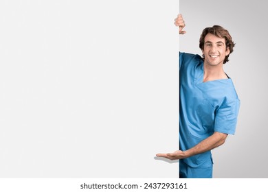 Smiling young male nurse in blue scrubs gesturing with one hand while playfully peeking from behind blank wall, ideal for interactive and engaging healthcare advertising 库存照片