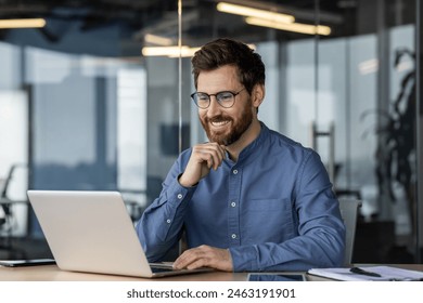 A smiling young man in glasses and a denim shirt is sitting in the office at a desk, working and talking on a video call online. Foto stock