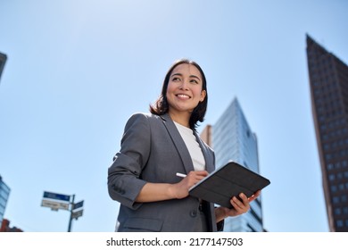 Smiling young Asian business woman leader entrepreneur, professional manager holding digital tablet computer using software applications standing on the street in big city on sky background. Stock-foto