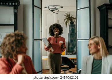 A smiling young black woman walks into an office with a laptop and a coffee to go, ready to have a meeting with her coworkers – Ảnh có sẵn
