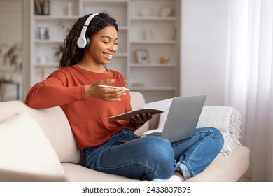 Smiling young black woman wearing headphones multitasking at home, writing in notebook while using laptop, happy african american female attending online course or watching webinar, sitting on couch Adlı Stok Fotoğraf