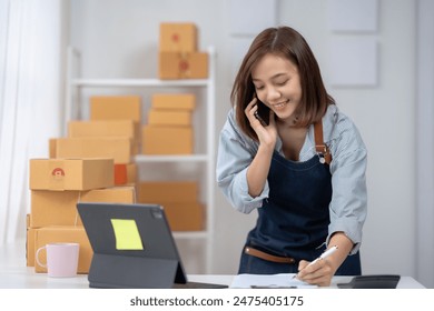 Smiling young woman managing a small business. She is on the phone taking orders and writing down notes, surrounded by packages. 库存照片