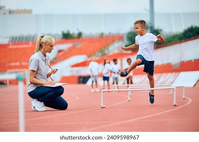 Sports teacher using stopwatch while boy is jumping over hurdles on running track. Focus is on kid.  Foto stock