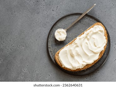 Spread cheese (ricotta, mascarpone, philadelphia or cream cheese) swirls on slice of oat wholegrain bread with tea spoon. Top view. Copy space. Grey background. Sandwich or toast on plate. ภาพถ่ายสต็อก