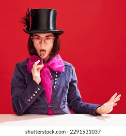 Stylish, attractive woman in cylinder hat and tailcoat with pink bow eating lollipop against red background. Confectionery factory boss. Concept of pop art, creativity, food, inspiration 库存照片