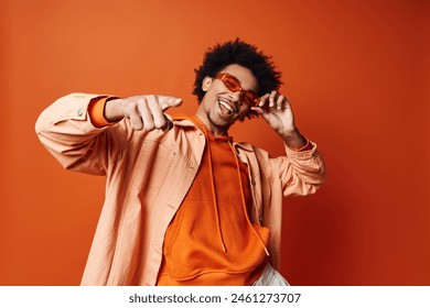 A stylish young African American man with curly hair wearing an orange shirt and sunglasses, posing energetically against an orange background. 库存照片