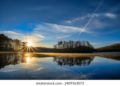 Sunrise at Chief McIntosh Lake in Indian Springs State Park, Georgia. The sun casts brilliant sunbeams while soft mist rises from the lake. The sky, clouds, and trees are reflected in the calm water. Foto stock