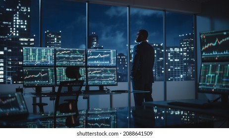 Successful Businessman Looking Out of the Window on Late Evening. Modern Hedge Fund Office with Computer with Multi-Monitor Workstation with Real-Time Stocks, Commodities and Exchange Market Charts. Foto stock