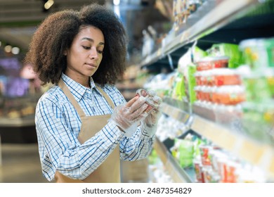スーパーの店員が食料品店の棚で商品ラベルを確認する。エプロンと手袋を着用して食品包装を調べる女性小売労働者。小売、食料品の買い物の品質管理のコンセプトの写真素材