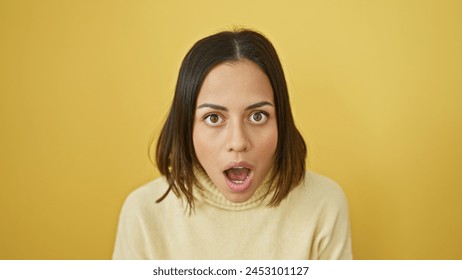 A surprised young hispanic woman with an open-mouth expression stands against a vibrant yellow background displaying intense emotion Foto Stock