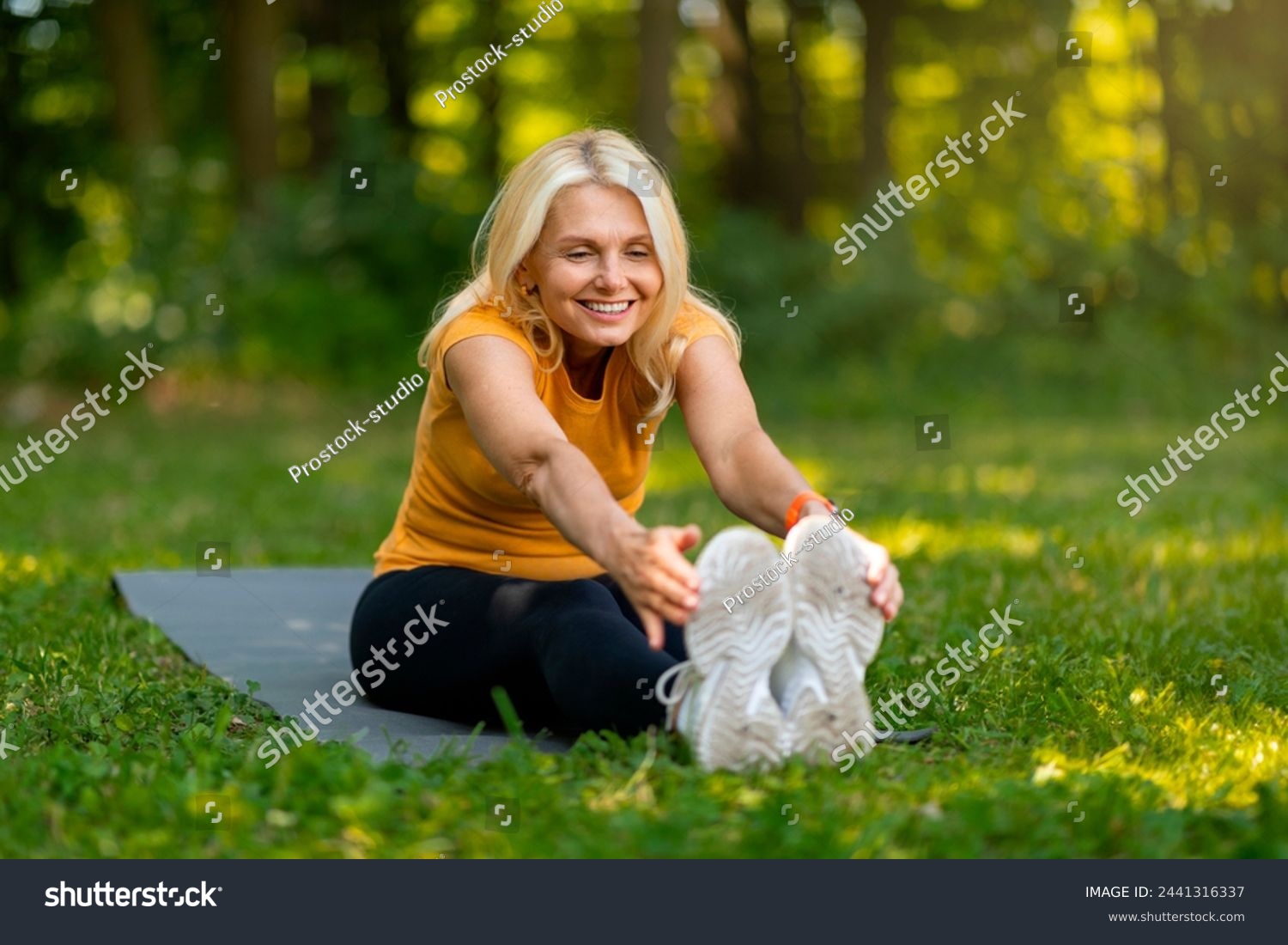Radiant senior woman with blonde hair stretching towards her toes on yoga mat in sunlit park, happy sporty elderly female training outdoors, exuding health and happiness, copy space