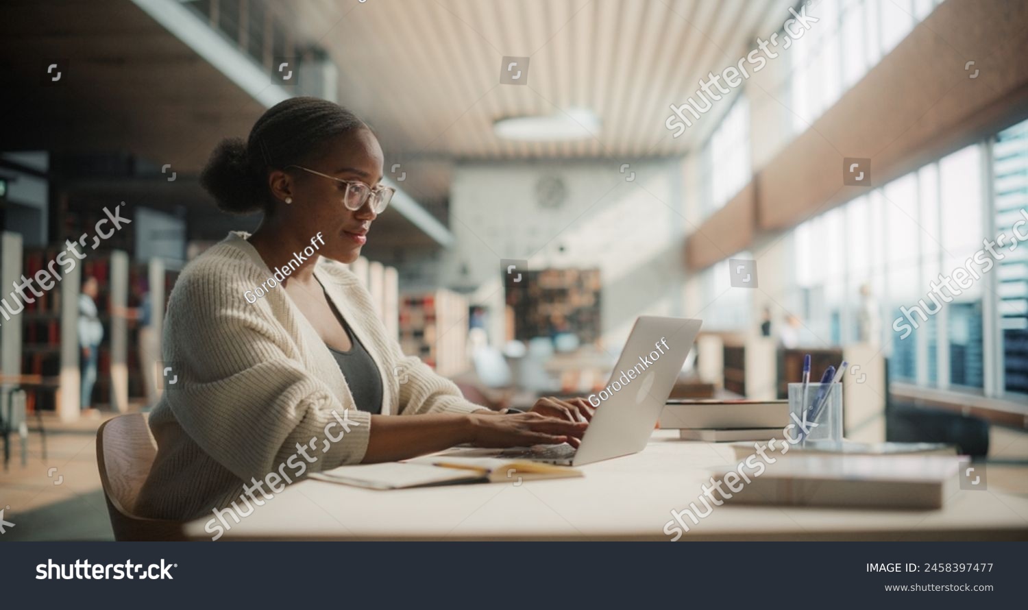 Dedicated African American Female Student Engaged in Online Learning at a Modern Library. Young Woman Using Laptop for Research, Surrounded by Books and Academic Environment.