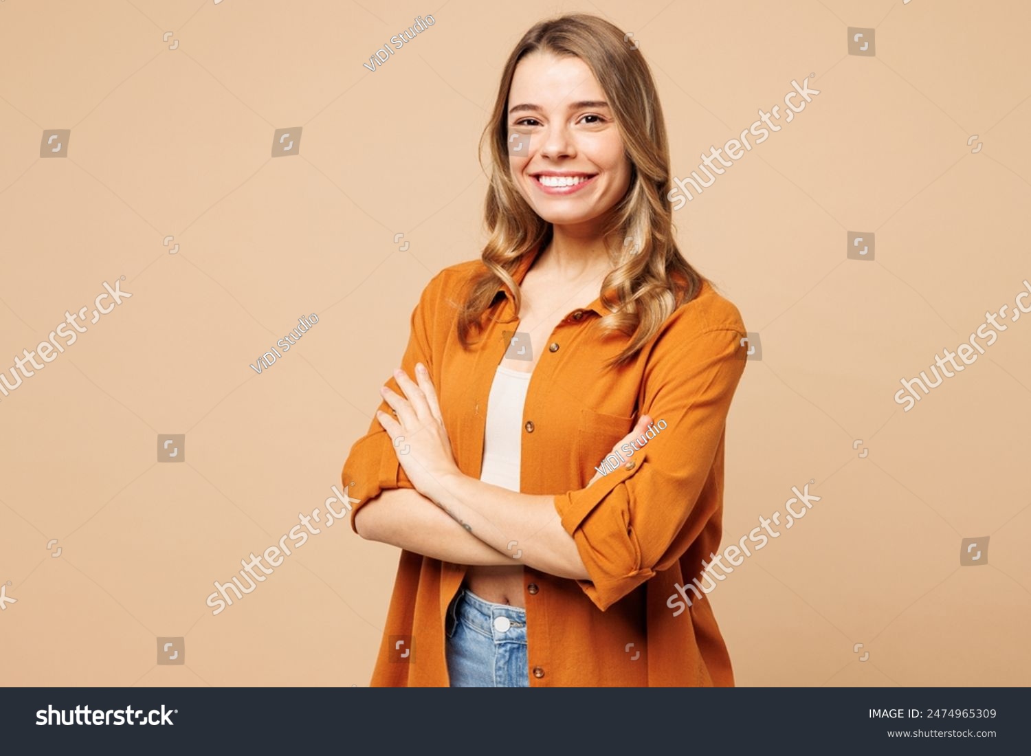 Vista lateral joven mujer caucásica sonriente que usa camisa naranja ropa casual sostenga las manos cruzadas cámara de mirada doblada aislada en liso pastel luz beige retrato de estudio de fondo. Concepto de estilo de vida