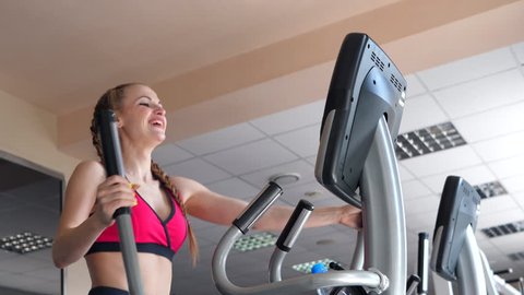 Sporty slim girl with long hair working out with the simulator in gym. She is engaged on a stationary bike. She diligently perform exercises and seeking plan. She is wearing in pink top. Slow motion Vídeo Stock