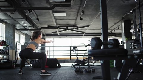 Tracking wide shot three quarter angle back view of athletic young woman in crop top and leggings doing squats to warm up in gym Video stock