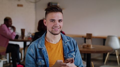 Young man with smartphone sitting in cafe, looking at camera. Stock-video