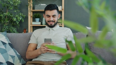 Carefree Arab man in casual clothing is using smartphone texting browsing indoors on couch at home. Modern devices, youth and communication concept. Stockvideo