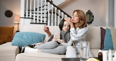 Beautiful Caucasian woman sitting on couch in living room with cute little sick daughter. Mother measuring temperature of her child. Unwell small girl with virus infection at home. Disease concept 库存视频