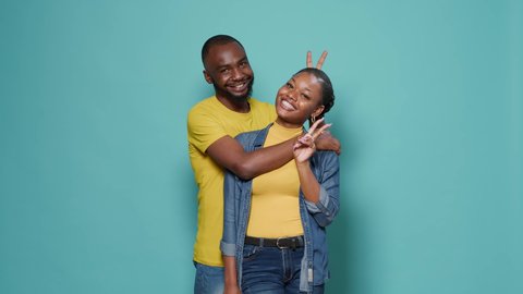 African american couple in embrace showing peace sign with hands, standing over blue background. Man and woman fooling around and advertising peaceful gesture together in studio. Video stock