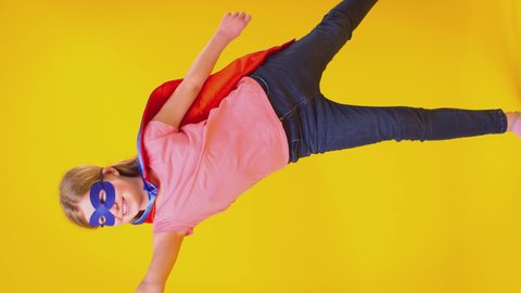 Vertical video of girl dressed as comic book super hero wearing mask and cape pretending to fly against yellow background - shot in slow motion Video stock