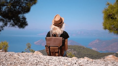 Girl with backpack and hat on top of mountain looking down on beautiful mountain valley with sea or ocean and islands raising her hands up admiring pace. Hiking. Nature.: stockvideo