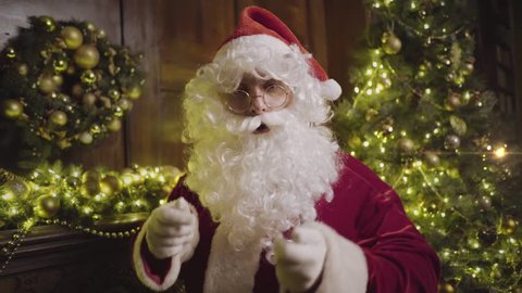 Santa Claus in eyeglasses blowing snow, sending kiss and looking at camera, decorated fireplace and christmas tree on background Stockvideo