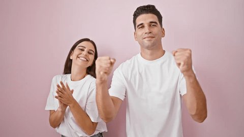 A cheerful young couple in white shirts clapping and smiling against a plain pink wall.の動画素材