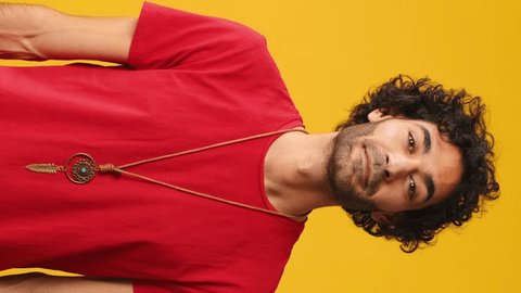 Vertical video, Surprised attractive man with curly hair, dressed in red T-shirt, covers his mouth with his palms in amazement looking at camera isolated on yellow background in studio : vidéo de stock