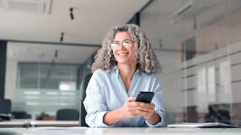 Happy middle aged business woman holding mobile cell phone using cellphone in office. Smiling mature older professional lady business owner entrepreneur using smartphone working sitting at desk. Arkistovideo