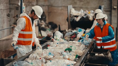 Shot from the side three workers in white uniforms in orange vests sort garbage near a conveyor belt transferring plastic bottles by color into the appropriate boxes at a waste recycling plantの動画素材