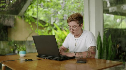 Transgender pro types on laptop in bright, plant-filled office. Engrossed in work, tattooed individual enjoys peaceful, productive environment. Inclusion, tech industry diversity represented. Slowmo Adlı Stok Video