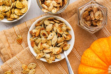 Bowl of toasted pumpkin seeds with a utensil, and in the surroudings, more toasted pumpkin seeds (with different toppings) in bowls and jars and an uncut pumpkin, all on light orange kitchen towel