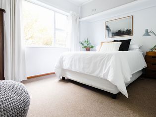 Bed with white sheets and neutral-colored pillows next to brightly-lit window on tan carpeted floor