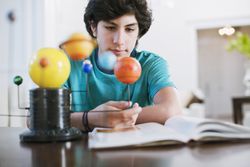 A high school student observes a model of the solar system while studying over a textbook